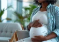 A pregnant woman sitting indoors with her hands resting on her belly.
