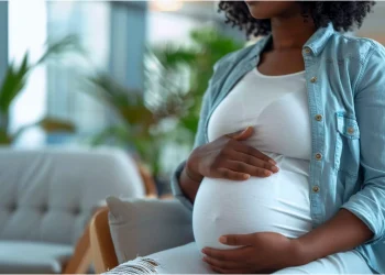 A pregnant woman sitting indoors with her hands resting on her belly.