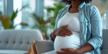A pregnant woman sitting indoors with her hands resting on her belly.